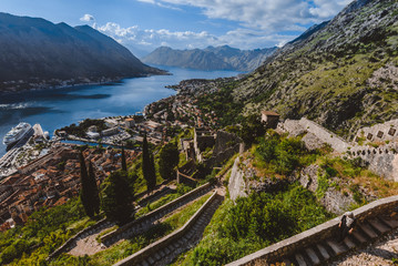 Fototapeta premium Kotor fortress walls and stone staircase wide angle view with Kotor Bay, Old Town rooftops and cloudy mountains on background. Boka Kotorska, Montenegro.