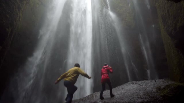 Young happy couple standing under beautiful waterfall Gljufrabui in Iceland and raises hands, feeling freedom and joy