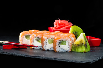Traditional Japanese cuisine. Selective focus on sweet sushi rolls with salmon, cream cheese, rice and kiwi on dark background, near ginger and kiwi