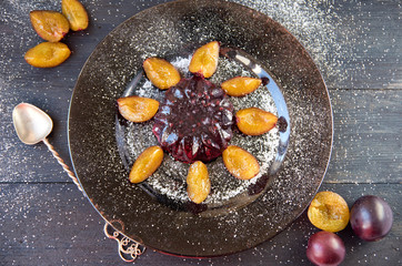 Cool purple jelly with fresh fruits and powder decorated with plums and silver vintage spoon on a dark brown plate on black wooden background. Top view