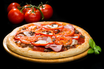 selective focus on appetizing Italian pizza with ham, tomato sauce, sausage, meat, cheese and sliced tomatoes on round wooden board on dark background, near tomatoes