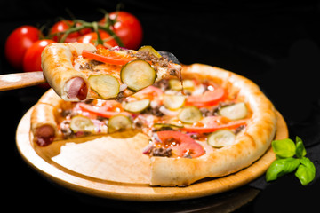 selective focus on sliced piece of appetizing Italian pizza with meat, onion, tomatoes, cheese and pickled cucumbers, dark background, close-up