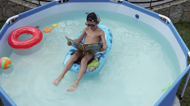 The boy reads a book and swims in the pool