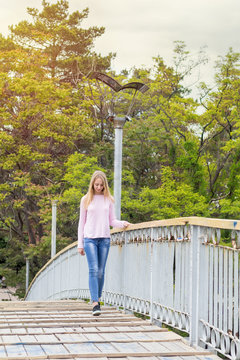 Blonde funny happy pretty girl in a pink sweatshirt on the bridge in park. Sunny day.