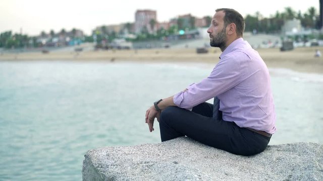 Pensive businessman admire view sitting on rock near sea
