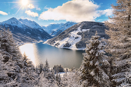 Lake and mountain range under cloudy sky with sun near Kaprun, Austria.