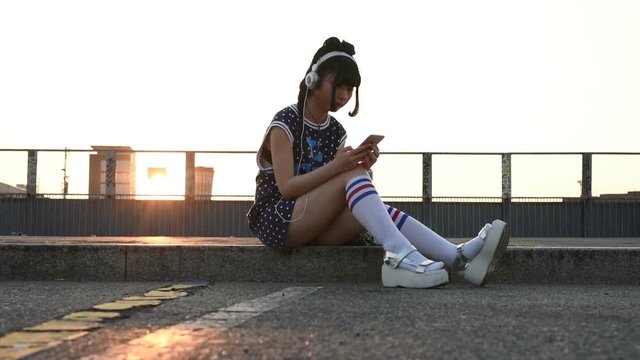 Young beautiful asian nonconformist woman listening music with headphones and smart phone hand hold outdoor in city back light, tapping the touchscreen - music, technology, social network concept