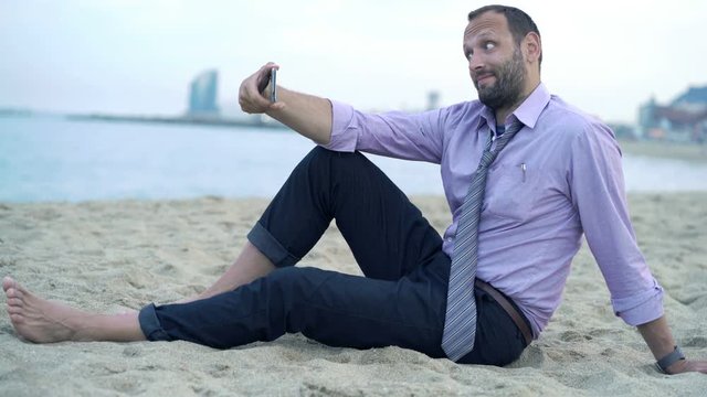 Happy businessman taking selfie photo with cellphone sitting on beach 

