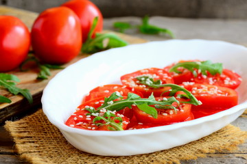 Ripe tomato slices, arugula rocket and sesame seeds salad. Garden tomato salad on a white plate and burlap textile. Dietary lunch or dinner idea. Closeup