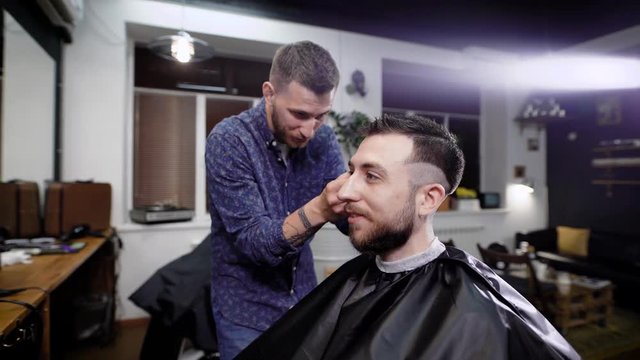 Close Up Of Stylish Smiling Bearded Man Sitting Covered With Black Peignoir In The Barbeshop. Barber Dressed In Casual Clothes Standing Behind Customer And Making Boxer Haircut With Clipper And Comb.