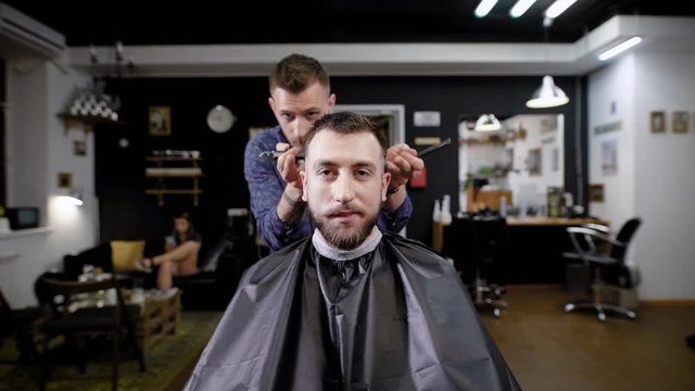Image of bearded hipster sitting in the barbershop covered with black peignoir. Barber dressed in casual clothes standing behind customer and making stylish haircut with hairclipper and comb.