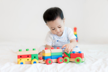 Adorable Asian baby boy 9 months sitting on bed and playing with color wooden train toys at home..