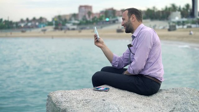 Young businessman watching movie on tablet computer sitting on rock near sea 

