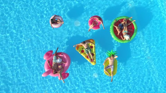 AERIAL TOP DOWN: Relaxed girls and guys laying on colourful floaties in pool. Happy smiling friends enjoying summer vacation on inflatable pineapple, pizza, flamingo, watermelon and doughnut floats