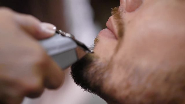 Image of face of bearded man getting grooming by professional hairdresser in modern barbershop. Close up of male chin and female hands using trimmer and getting perfect shape for beard and moustache.