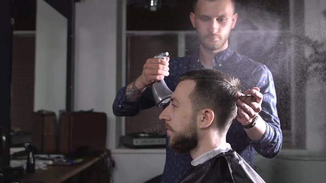 Image of stylish male barber spraying water on hair of client and brushing it to make stylish haircut. Young hairdresser working at the barbershop doing undercut with comb and styling products.