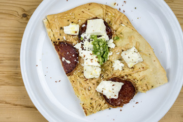 Slice of homemade salami pizza with feta cheese and seasoning served on a white plate on wood