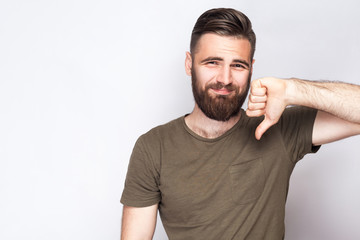 Portrait of unsatisfied bearded man with thumbs down and dark green t shirt against light gray background. studio shot.  .