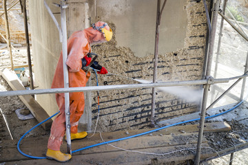 Worker in orange protective suit cleans corrosion damaged concrete bridge pillar with high pressure washer