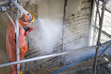 Worker in orange protective suit cleans corrosion damaged concrete bridge pillar with high pressure washer