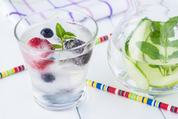 Glass of cucumber, mint infused water with ice cubes with fresh berries and pitcher.