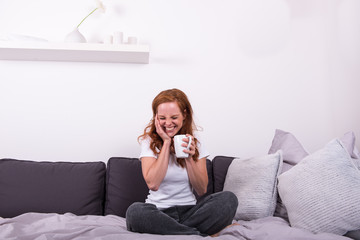 junge, rothaarige Frau sitzt auf dem Bett und freut sich über Ihren Kaffe