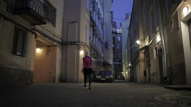 shortcut blondie woman goes out from inner yand to the street at dark night. There are electric lights on buildings. She wears black dress, pink jacket, clutch bag, Saint Peterburg, Russia
