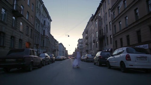 shortcut blondie woman in white dress is going and dancing the street at early morning before sunrise. There are cars parking on both side of the street and perspective of buildings. Saint Peterburg, 