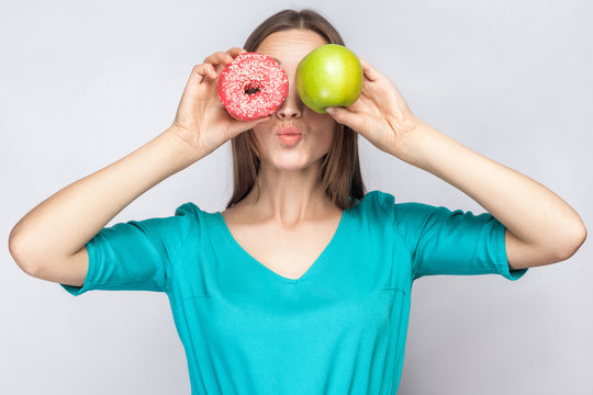 Beautiful young woman with freckles in green dress, holding before her eyes green apple and pink donut and kissing. studio shot on light gray background.