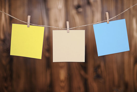 Close up of three yellow beige and blue note papers hung by wooden clothes pegs on a brown wooden background