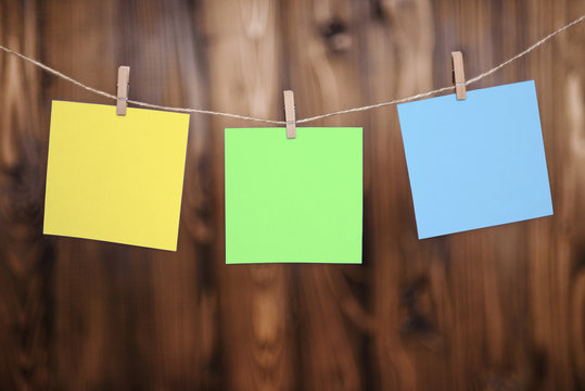 Close up of three yellow green and blue note papers hung by wooden clothes pegs on a brown wooden background