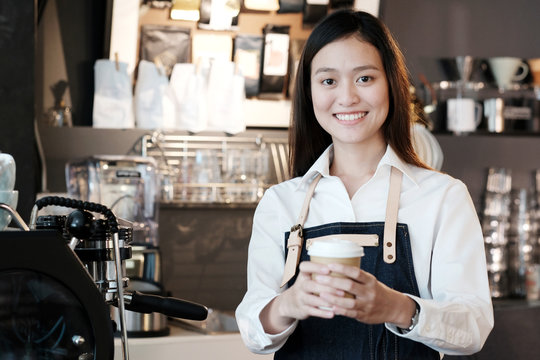 Young asian women Barista holding a take away coffee cup with smiling face at cafe counter background, small business owner, food and drink industry concept