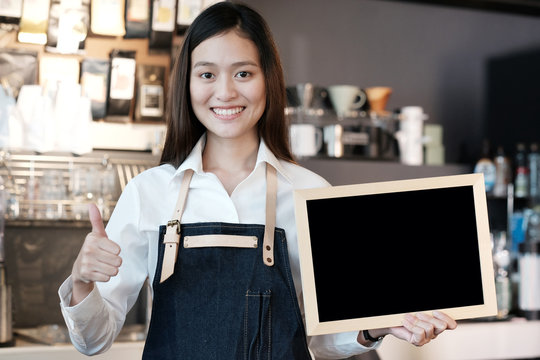 Young asian women Barista holding blank chalkboard and thumb up gesture with smiling face in font of cafe counter background, small business owner, food and drink industry concept