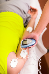 Close-up of a personal trainer holding with one hand the electric machine with the electrostimulator electrodes in the arm of a deportist, wearing a green t-shirt fluorescent, upside down on a bed