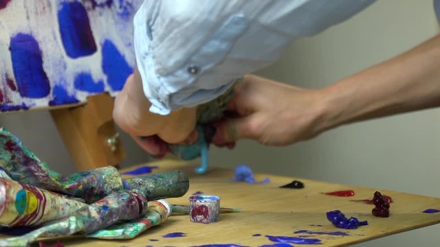 The girl squeezes blue oil paint on a wooden palette full oh paint tubes, slow motion shooting