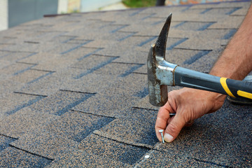 Worker hands installing bitumen roof shingles. Worker Hammer in Nails on the Roof. Roofer is hammering a Nail in the Roof Shingles. Unfinished roof. © cherokee4