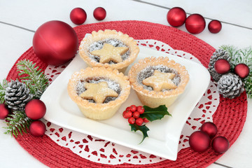 Homemade christmas mince pies on a plate with holly, fir and  red bauble decorations on a place mat on distressed white wood background.