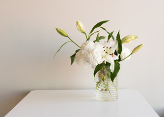 Bouquet of white flowers, chrysanthemums and lilies in glass jar on white table against neutral wall background