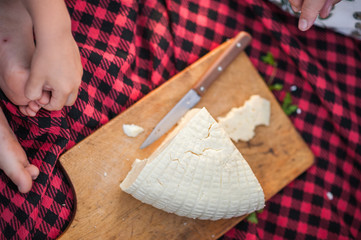 Hands hold a knife. The baby's hands cut the cheese. A child cuts goat cheese on a kitchen board