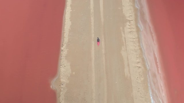 AERIAL CLOSE UP: Unrecognizable female walking along the sandy levee surrounded by gorgeous orange water of Las Colorado saltworks on hot sunny day in Mexico. Tourist girl strolling along salt ponds