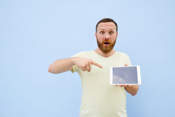 Upset Handsome young man with a beard showing something on a tablet isolated on a blue background, bright and light