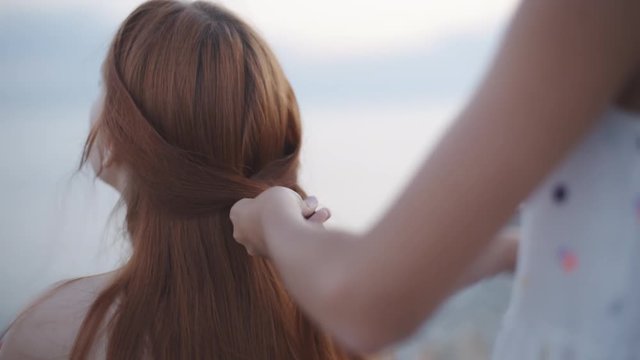 Anonymous woman standing behind redhead girl and braiding her hair on beach in sunlight.