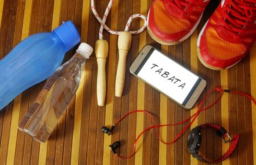 Fitness, healthy and active lifestyles concept. Dumbbells, sport shoes, smart phone with earphone, skipping rope and water bottle gym floor with word tabata