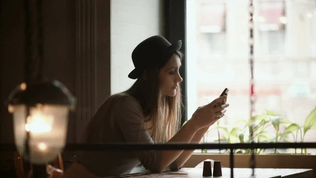 Stylish young woman in hat using the smartphone in cafe sitting near the window. Attractive female surfing the Internet.