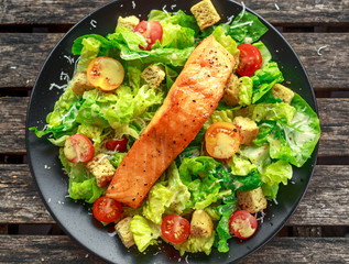 Caesar Salmon Salad with croutons, parmesan cheese, tomatoes, dressing and pepper in a black plate on wooden table.