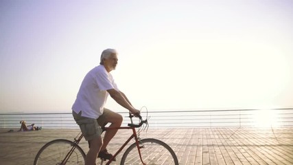 Handsome senior man riding bike on seafront, slow motion