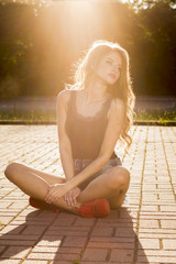 Fashionable blonde model dressed in ripped jeans shorts and black shirt sitting on the paving tiles at sunset