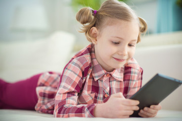 Pretty little child girl with digital tablet, looking and smiling while lying on white couch