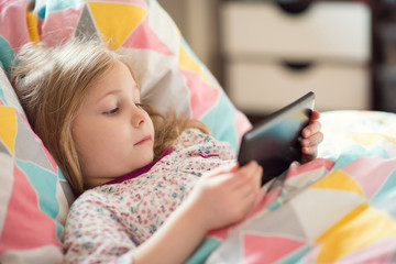Pretty little child girl with digital tablet, looking and smiling while lying in bed