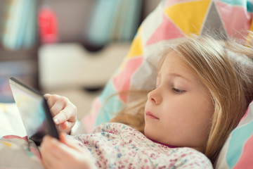 Pretty little child girl with digital tablet, looking and smiling while lying in bed
